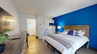 This well-appointed single-floor unit offers a smart, space-conscious layout that combines the sleeping and living areas into one cohesive open space. The main room is immediately distinguished by a striking cobalt blue accent wall, which frames the sleeping area and provides a vibrant focal point against the otherwise neutral white walls and light wood-look flooring that runs throughout the unit.

The kitchenette is neatly integrated along one wall, featuring white cabinetry, a compact refrigerator, microwave, and countertop cooking surface — providing essential functionality without compromising the open feel of the space. A wall-mounted flat-screen television and a wood-toned dresser with ample drawer storage add practicality to the room, while a sliding glass door with vertical blinds allows natural light to filter in and provides access to an outdoor area.

The unit includes two bathrooms, one of which is visible as a bright, white-finished space with vanity cabinetry and mirror, and a second featuring a full bathtub with shower. A dedicated closet area with wire shelving and hanging rods offers organized storage. The overall design is clean and functional, with a modern aesthetic punctuated by warm wood tones and bold color choices that give the space a distinct, welcoming character.