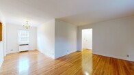 Located at Knickerbocker Rd in Dumont, NJ, this well-appointed one-bedroom, one-bathroom unit offers a practical and comfortable layout across a single floor. Gleaming hardwood floors run throughout the living areas, bedroom, and dining room, lending warmth and continuity to the space. The living room is open and airy with clean white walls, providing a versatile canvas for furnishings. A pass-through window connects the dining room to the kitchen, enhancing the sense of openness. The dining room is anchored by a classic chandelier and benefits from natural light through a window with a view of greenery outside, complemented by a wall-mounted radiator. The kitchen is equipped with warm oak cabinetry, dark countertops, and a full suite of stainless steel appliances including a gas range, over-the-range microwave, refrigerator, and dishwasher. The bedroom features hardwood floors, a wall-mounted air conditioning unit, a radiator, and a bright window overlooking the outdoors. The bathroom is cleanly finished with marble-style tile flooring, a white vanity, and a glass-enclosed walk-in shower with a window for natural ventilation. Crown molding details are present throughout, adding a refined touch to the overall design.