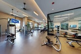 This well-appointed fitness center is designed to accommodate a comprehensive range of workout activities across several distinct zones. The main cardio area features a generous row of treadmills, ellipticals, and stationary bikes, all positioned near large, dark-framed windows that bring in ample natural light and provide outdoor views. Ceiling fans and recessed linear lighting maintain a bright, comfortable atmosphere throughout.

A floor-to-ceiling glass partition elegantly separates the main floor from a dedicated studio space, which includes hardwood flooring, wall-mounted mirrors, a ballet barre, and an organized dumbbell rack with a full range of free weights. This studio is well-suited for group fitness classes, stretching, or individual training sessions. Mounted flat-screen televisions are positioned throughout the facility for entertainment during workouts.

The strength training area houses a variety of cable machines and resistance equipment, with open floor space in between for functional movements. Near the entrance, a built-in refreshment station with a water dispenser and small refrigerator is set against a rich wood-paneled accent wall, offering a polished, hotel-quality finish. The overall design is modern and cohesive, with neutral tones and high-end finishes throughout.
