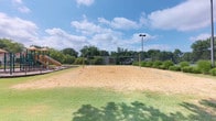 This outdoor recreational space is designed to encourage active play and community engagement. At its center sits a regulation-style sand volleyball court, complete with a net strung between two upright poles and clearly marked boundary lines. The court's sandy surface provides an ideal playing environment for both casual and competitive matches. A dedicated light pole stands adjacent to the court, suggesting the space can accommodate evening use. To the left of the volleyball court, a children's playground is visible, featuring at least one slide structure, making this area suitable for a range of ages. The surrounding landscape is well-kept, with neatly trimmed grass extending outward from the court and mature deciduous trees lining the perimeter, providing a natural green backdrop. Low ornamental shrubs are planted along the edges, adding definition to the space. The open-sky setting allows for ample natural light throughout the day. The overall layout is functional and spacious, offering room for spectators and additional recreational activities alongside the main court.