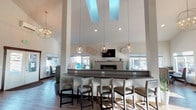 Located at 9118 East Columbia Drive in Spokane, Washington, this single-floor leasing office for Northwood Apartments is designed to impress and inform prospective residents. The centerpiece of the space is a dramatic open lobby featuring soaring vaulted ceilings with large skylights that flood the interior with abundant natural light. A substantial reception bar with a dark countertop and upholstered bar seating serves as the focal point, complemented by globe pendant lights and a geometric chandelier that add a modern, refined touch. Behind the bar, a built-in fireplace and wall-mounted television enhance the welcoming atmosphere, while large windows on multiple sides provide views of the surrounding exterior grounds. The entryway includes a comfortable lounge seating area with accent chairs and a side table, positioned beside windows that bring in additional daylight. A branded interior window with the Northwood Apartments logo adds a polished, professional identity to the reception zone. A private conference room off the main area offers a dark-topped meeting table with upholstered seating and stylish pendant lighting, ideal for one-on-one leasing consultations. A compact kitchenette is also present, equipped with a stainless steel refrigerator, microwave, sink, and coffee station. Wood-look flooring flows throughout, tying together the contemporary and functional design of this well-appointed leasing office.