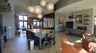 This single-floor space showcases a thoughtfully designed open-plan interior that blends modern industrial and rustic elements seamlessly. At the heart of the room sits a substantial concrete-topped communal table surrounded by metal industrial-style chairs, illuminated dramatically by an eye-catching cluster of large globe pendant lights with geometric patterned shades. The herringbone-patterned wood-look flooring adds warmth and visual texture underfoot, complementing the overall aesthetic.

A prominent stone fireplace anchors one wall, providing a cozy focal point and serving as a natural gathering area. Nearby, a built-in bar-height counter with bar stools offers an additional seating option. The space is further enriched by a striking decorative installation featuring birch logs and reclaimed dark wood paneling, adding an organic, nature-inspired contrast to the contemporary finishes.

Large windows along the perimeter allow abundant natural light to flow through the space, enhancing its welcoming atmosphere. Accent furnishings, including an orange upholstered chair, introduce warm color tones. The combination of curated décor, varied seating arrangements, and layered lighting creates an inviting and versatile communal environment.
