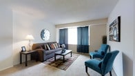 This single-floor space offers a thoughtfully designed layout centered around a welcoming living area. The living room serves as the focal point of the space, featuring plush wall-to-wall carpeting in a warm neutral tone that creates a soft, inviting foundation throughout. Large windows dressed with deep navy-blue curtains allow natural light to filter in while adding a rich contrast to the light beige walls, contributing to a calm and balanced atmosphere.

The living area is furnished with a generously sized gray sofa accented with decorative throw pillows, complemented by a pair of bold teal accent chairs that introduce a pop of color and personality to the room. A dark-finished rectangular coffee table anchors the seating arrangement, set atop a geometric-patterned area rug that ties the color palette together.

A decorative circular mirror mounted on the wall adds a stylish artistic element, while a classic table lamp provides warm ambient lighting. The textured ceiling adds subtle architectural character to the space. Overall, the space presents a clean, modern aesthetic with a harmonious blend of neutral and accent tones, making it a comfortable and visually appealing environment suited for both relaxing and entertaining.