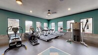 Located at 6515 W Hausman Rd in San Antonio, Texas, this fitness center is a well-equipped, single-floor facility designed to support a comprehensive workout experience. The space is characterized by bold teal accent walls, a reflective diamond-plate wainscoting detail, and a combination of durable rubber flooring and a colorful geometric-patterned carpet in the central exercise area.

Natural light fills the room through a generous arrangement of large windows along multiple walls, offering views of the surrounding community. Recessed lighting and a ceiling fan provide additional illumination and air circulation throughout the space.

The cardio zone features multiple treadmills and elliptical machines, all equipped with individual digital displays. A cable pulley strength machine and a multi-function weight station serve users focused on resistance training, while a rack of dumbbells and a selection of exercise balls cater to free-weight and functional fitness routines.

Two wall-mounted flat-screen televisions are positioned for easy viewing during workouts. A mirrored wall section expands the sense of space and supports form monitoring during exercise. A vending machine stocked with snacks and beverages provides convenient on-site refreshment. The overall design is energetic and functional, making this fitness center a practical and inviting amenity.