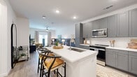 This thoughtfully designed single-floor unit offers an efficient and modern living experience within a compact footprint. The open-concept kitchen and living area serves as the heart of the space, seamlessly blending functionality with contemporary style. The kitchen is outfitted with grey shaker-style cabinetry, stainless steel appliances including a French door refrigerator, range, microwave, and dishwasher, complemented by a herringbone tile backsplash and a white quartz waterfall island with an undermount sink and bar seating for two. Light wood-look flooring runs throughout, enhancing the bright, airy atmosphere created by recessed lighting overhead.

The living area features a distinctive two-tone wall treatment with a deep slate-blue lower half, adding a modern design accent. A ceiling fan, large window with drape treatments, and access to a private balcony bring natural light and outdoor connectivity to the space. The balcony overlooks a landscaped green area with neighborhood views.

The bathroom is clean and well-appointed, featuring grey vanity cabinetry matching the kitchen, a tub-shower combination with white tile surround, and light wood-look flooring. A hallway and dedicated closet provide practical storage solutions, completing this efficiently organized unit.