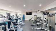 This fitness center offers a spacious, single-floor exercise facility designed to support a wide range of workout routines. The room is finished with neutral, light-toned walls and durable rubber flooring, creating a clean and professional environment suited for both casual and dedicated fitness use.

The cardio zone is prominently featured along one wall, lined with a row of Precor elliptical machines and treadmills. Large multi-pane windows allow abundant natural light to fill this area, providing views of surrounding greenery and contributing to an open, energizing atmosphere.

The strength-training section is equally well-appointed, featuring cable and weight machines, adjustable benches, and a full dumbbell rack with a broad range of weights. Full-length mirrors are mounted along the walls, enhancing the sense of space while allowing users to monitor their form. A wall-mounted flat-screen television is positioned for visibility across the workout area.

Recessed lighting throughout the ceiling ensures consistent illumination during all hours of use. The layout is thoughtfully organized to allow comfortable movement between equipment zones. The facility also includes a dedicated entryway and an adjacent hallway, suggesting access to additional amenities nearby. Overall, this fitness center is a comprehensive, professionally outfitted space built to serve a broad range of exercise needs.