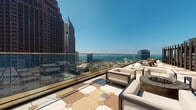 Located along Euclid Avenue in the heart of downtown Cleveland, this single-floor amenity space is designed to offer a sophisticated and comfortable environment with striking panoramic views of the city skyline and Lake Erie. Floor-to-ceiling windows line the interior, flooding the space with abundant natural light and providing unobstructed sightlines toward the waterfront and surrounding urban landscape.

The open-concept layout seamlessly connects several distinct functional zones. A fully equipped kitchen anchors one end of the space, featuring stainless steel appliances, a double oven range, integrated dishwasher, and sleek gray cabinetry with white countertops set atop warm hardwood flooring. Textured wall treatments add visual depth to the kitchen area.

Adjacent to the kitchen, a dining and co-working zone is furnished with high-top tables and bold burgundy and gold bar chairs, positioned directly in front of floor-to-ceiling sliding glass doors that open onto the outdoor terrace.

The lounge area is warmly appointed with a leather sofa, colorful accent seating, a built-in bookshelf, and a large television, creating an inviting, residential atmosphere. The expansive outdoor terrace features glass railings, patterned stone tile, and comfortable lounge furniture, offering an exceptional setting to enjoy sweeping views of Cleveland's skyline and lakefront.