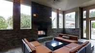 This covered outdoor living space makes a striking impression with its soaring stacked stone fireplace as the undisputed focal point. The fireplace rises from a slate-toned hearth to the ceiling, flanked by wall-mounted lantern sconces that cast a warm, inviting glow. The stacked stone cladding extends across the surrounding walls, creating a cohesive, rustic-yet-refined aesthetic throughout the space.

Large windows line multiple walls, flooding the interior with abundant natural light while framing lush green views of the surrounding landscape. The white paneled ceiling adds a crisp contrast to the rich, dark stonework below, enhancing the sense of openness and height.

The lounge area is furnished with generous modular seating upholstered in warm cognac leather, arranged around a sleek dark-framed coffee table with a reflective top — ideal for gathering and entertaining. Decorative accent pillows add subtle texture to the seating arrangement.

The flooring features cool-toned stone tile, complementing the natural materials used throughout. A ceiling fan overhead ensures comfortable airflow, making this space enjoyable across multiple seasons. Overall, the area combines rustic architectural character with modern furnishings to create a welcoming, functional outdoor retreat.