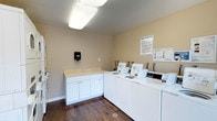 This shared laundry facility is designed with functionality and convenience in mind. The room features a row of commercial-grade top-loading washing machines and dryers neatly arranged along one wall, providing ample capacity for residents. Adjacent to the machines is a built-in white cabinetry unit topped with a light-colored laminate counter, offering a dedicated space for folding laundry or sorting clothes. A wall-mounted sanitary dispenser is conveniently positioned nearby for user hygiene. The room is finished with warm beige walls and rich dark hardwood-style flooring, creating a clean and welcoming environment. Overhead, flush-mounted fluorescent light fixtures provide bright, even illumination throughout the space. Laundry facility rules are clearly posted on the wall, ensuring organized and respectful use of the shared amenities. The layout is open and practical, allowing for easy movement between machines and the folding station. Overall, this laundry room is thoughtfully outfitted to serve the needs of multiple users efficiently and comfortably.