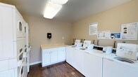 This shared laundry facility is designed with functionality and convenience in mind. The room features a row of commercial-grade top-loading washing machines and dryers neatly arranged along one wall, providing ample capacity for residents. Adjacent to the machines is a built-in white cabinetry unit topped with a light-colored laminate counter, offering a dedicated space for folding laundry or sorting clothes. A wall-mounted sanitary dispenser is conveniently positioned nearby for user hygiene. The room is finished with warm beige walls and rich dark hardwood-style flooring, creating a clean and welcoming environment. Overhead, flush-mounted fluorescent light fixtures provide bright, even illumination throughout the space. Laundry facility rules are clearly posted on the wall, ensuring organized and respectful use of the shared amenities. The layout is open and practical, allowing for easy movement between machines and the folding station. Overall, this laundry room is thoughtfully outfitted to serve the needs of multiple users efficiently and comfortably.