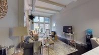 This two-storey space showcases a compelling blend of industrial and contemporary design elements. The main living area immediately draws the eye upward to its soaring ceiling, where exposed structural beams and a prominent metallic duct run overhead, lending the space an authentic loft-like character. Large, multi-pane windows along the exterior wall invite abundant natural light, offering views of the surrounding urban streetscape and creating an airy, open atmosphere throughout.

The living area is generously proportioned, accommodating a full seating arrangement that includes a tufted leather sofa, accent chairs, and a layered area rug anchoring the space. Decorative touches such as hanging planters, metallic side tables, and ornate console furnishings add warmth and contrast to the raw architectural elements. Dark hardwood flooring runs throughout, grounding the cool grey wall tones and white trim. A staircase with sleek metal railings connects to the second floor, reinforcing the space's modern-industrial aesthetic. The overall ambiance balances sophistication with an approachable, lived-in comfort suited to urban living.