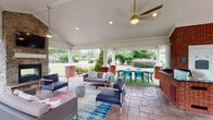 Located at 2000 Geddy Ln in Cary, NC, this generously proportioned covered outdoor pavilion offers a well-appointed gathering space designed for both relaxation and entertainment. The structure features a vaulted white shiplap ceiling supported by classic white columns rising from brick bases, lending the space a polished, traditional character. Ceiling fans with stylish gold fixtures are positioned throughout, ensuring comfortable airflow year-round.

The stamped concrete flooring provides a durable yet refined foundation across the entire pavilion. A stone fireplace anchors one end of the space, creating a cozy focal point alongside a seating arrangement of contemporary navy blue armchairs and sofas accented by colorful area rugs. Adjacent to the lounge area, a dedicated outdoor kitchen station features a built-in grill and preparation surfaces framed in classic brick, complemented by a separate dining area furnished with a dark table and vibrant teal chairs set on artificial turf.

Open on multiple sides, the pavilion offers unobstructed views of the surrounding lush landscaping, including manicured shrubs, flowering plants, and mature trees. Natural light fills the space freely, while recessed lighting and pendant fixtures ensure usability into the evening hours. The overall ambiance is inviting, functional, and thoughtfully designed for outdoor living.