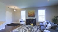 This single-floor living space offers a thoughtfully designed interior centered around a comfortable and stylish living area. The room is characterized by neutral gray walls and smooth ceilings, creating a calm, contemporary backdrop. A pendant light fixture hangs overhead, adding a subtle decorative touch to the space.

Natural light flows in generously through multiple double-hung windows fitted with white blinds, brightening the room throughout the day. The living area features a plush gray sofa accented with patterned throw pillows, paired with a sleek acrylic-legged accent chair adorned with a decorative "smile" pillow, adding a playful yet chic element to the setting.

A bold geometric-patterned area rug in gray and white anchors the seating arrangement, complemented by a glass and metal coffee table at the center. A dark wood media console serves as a focal point along one wall, topped with decorative objects and a vibrant warm-toned canvas artwork depicting bare branches — providing a striking contrast against the neutral surroundings.

The overall ambiance is modern and welcoming, blending clean lines with carefully curated accessories for a polished, livable aesthetic.