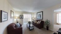 This single-floor residential unit offers a thoughtfully designed layout that balances comfort and functionality across its key living spaces. The living room serves as the heart of the home, featuring soft neutral walls, plush wall-to-wall carpeting, and a large sliding glass door dressed with vertical blinds that fills the room with natural light and provides access to an outdoor area. The open, airy feel is complemented by ample space for seating and entertaining. Adjacent to the living room, the kitchen is efficiently designed with white cabinetry, a coil electric range, a stainless steel double sink, and a pass-through breakfast bar with bar stool seating, connecting seamlessly to the dining area. The dining space accommodates a full table and chairs and features warm neutral tones with a window for additional natural light. The bedroom is generously proportioned with plush carpeting, a large window with drapes, and direct access to a walk-in closet, offering practical storage. The full bathroom includes a vanity with under-sink storage, a toilet, and a shower-tub combination with tile surround and hardwood-style flooring, providing a clean and functional finish. Throughout the unit, warm beige tones and consistent finishes create a cohesive, welcoming ambiance.