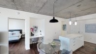 This single-floor space showcases an industrial-modern aesthetic defined by its striking exposed concrete ceiling, which serves as a dramatic architectural focal point throughout. Dark hardwood flooring grounds the interior, creating a warm contrast against the crisp white walls and cabinetry.

The open-plan layout integrates a dining area anchored by a round glass-top table surrounded by contemporary upholstered chairs. A bold black dome pendant light hangs overhead, complementing the industrial character of the ceiling. Adjacent to the dining area, a white kitchen island with drawer storage provides additional workspace and casual seating potential.

A built-in desk nook is seamlessly integrated into the white walls, offering a dedicated workspace complete with shelving and task lighting—an efficient use of the available space. The nook is decorated with a monochromatic art print and a vibrant orchid, adding personality to the area.

Doorways lead to additional rooms beyond, suggesting a well-connected floor plan. A second pendant light and colorful wall art are visible in a nearby corridor or room, reinforcing the space's cohesive contemporary style. Overall, the design balances urban industrial elements with clean, modern finishes.