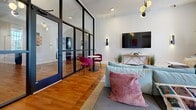 This 730 square foot clubroom, located at 2110 Pickens St in Columbia, SC, offers a contemporary and multifunctional space. The single-floor layout is characterized by its modern architectural elements, most notably the sleek glass partitions that create distinct areas while maintaining an open feel. Rich hardwood flooring runs throughout, adding warmth to the contemporary design.

The space is divided into several functional zones. A lounge area features a comfortable light blue sofa adorned with colorful throw pillows, perfect for relaxation or casual meetings. Adjacent to this is a substantial wooden table, ideal for group work or dining. Large windows dressed with vibrant yellow curtains flood the room with natural light, enhancing the airy atmosphere.

A separate section, enclosed by the glass partitions, houses a small seating area with bold fuchsia chairs and a round table, suitable for more private conversations or focused work. The room is further equipped with modern amenities including a wall-mounted large-screen TV and stylish lighting fixtures that blend form with function.

The decor strikes a balance between professionalism and comfort, with touches of greenery and abstract wall art adding visual interest. The color palette, dominated by whites and woods with pops of bright colors, creates an energetic yet welcoming environment. This versatile clubroom is designed to accommodate various activities, from casual socializing to professional meetings, all within a fresh and inviting atmosphere.