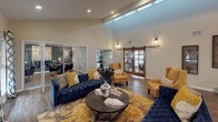 This well-appointed single-story communal space is thoughtfully designed to accommodate both social gatherings and private meetings. The centerpiece is a generously proportioned lounge area distinguished by soaring vaulted ceilings that lend an open, airy feel. Dramatic vaulted ceilings draw the eye upward, while recessed lighting and clerestory windows allow natural light to filter throughout. The lounge features a wood-accented tiled fireplace with a wall-mounted television above it, flanked by large windows dressed in bold geometric-patterned drapes. Deep navy tufted sofas, mustard accent chairs, and a patterned area rug create a cohesive, contemporary aesthetic.

Glass-panel sliding doors divide the lounge from an adjacent dining area, which features a rustic wood dining table beneath a striking multi-globe sputnik chandelier. A second, more intimate dining or meeting space offers a round table set against a slate-blue accent wall. A rustic barn door leads to a dedicated workspace or office area, adding a distinctive architectural detail.

The galley kitchen is outfitted with gray cabinetry, granite-style countertops, and dark appliances, providing practical functionality. Throughout the space, wood-look flooring ties each area together, contributing to a warm, modern, and inviting ambiance.
