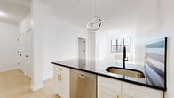 This thoughtfully designed one-bedroom, one-bathroom residence at 540 Bond Street offers a clean, modern aesthetic defined by crisp white walls, light hardwood flooring, and bold black hardware accents throughout. The open-concept kitchen and living area serves as the heart of the unit, anchored by a central island with striking dark countertops, white shaker cabinetry with gold pulls, a stainless steel dishwasher, and a full suite of stainless steel appliances including a refrigerator, oven, and over-the-range microwave. A sculptural loop pendant light adds a contemporary focal point above the island. The living area benefits from large black-framed windows that invite abundant natural light and offer views of the surrounding neighborhood. The bedroom is a bright, serene retreat, featuring generous closet space with multiple built-in closets finished with matching black hardware, and a large window that bathes the room in sunlight. The bathroom is finished with terrazzo-style tile flooring, a soaking tub, and matte black fixtures, maintaining the unit's cohesive modern palette. Four closets provide ample storage throughout. The overall ambiance is sleek, minimalist, and move-in ready.
