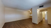 This single-floor space showcases an open-concept layout that seamlessly connects the main living area to the kitchen. The living area features warm, dark wood-style laminate flooring that extends throughout, providing a cohesive and grounded aesthetic. The textured popcorn ceiling adds a classic touch, while white baseboards offer a clean contrast to the flooring below.

The living space is generously proportioned, offering a blank canvas with neutral beige walls that provide flexibility for various furniture arrangements and décor styles. A ceiling fan is centrally positioned in the kitchen area, ensuring air circulation throughout the connected spaces.

The kitchen, visible from the main living area, features light wood cabinetry and a countertop with a sink, benefiting from the open sightlines that make the overall space feel larger. A wall-mounted sconce near the entryway door adds a warm, functional lighting element, complementing the natural and overhead light sources.

The entryway is framed by a clean doorway with a wall sconce, creating a welcoming transition into the space. Overall, the unit presents a straightforward, functional layout with a neutral palette suited for comfortable everyday living.