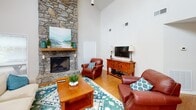 This single-story space is thoughtfully organized into distinct functional zones, each with its own character and purpose. The living area serves as the heart of the space, commanding immediate attention with its soaring vaulted ceilings and a striking floor-to-ceiling stone fireplace that anchors the room. Rich leather seating, warm hardwood flooring, and a patterned area rug in teal and ivory tones create an inviting, comfortable atmosphere. Artwork displayed above the stone mantle adds a decorative focal point to the dramatic architectural feature.

Adjacent to the lounge area, a generously sized dining space accommodates multiple round tables paired with industrial-style metal chairs, making it well-suited for group gatherings. Vaulted ceilings continue here, and an abundance of windows fitted with horizontal blinds allows natural light to fill the room. A pendant light fixture adds a refined touch overhead. A dedicated seating nook near an exterior door provides additional casual dining space with views of the surrounding exterior.

The kitchen features white cabinetry, bold dark granite countertops with dramatic veining, and a large bar-height island ideal for casual seating. Beadboard backsplash detailing and track lighting lend a classic yet functional character to this well-equipped area.