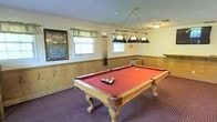 The game room at Mission Viejo Villas offers a welcoming recreational space designed for social gatherings and leisurely play. At the center of the room stands a full-size pool table with a bold red felt surface and elegant carved wooden legs, positioned beneath a classic brass hanging light fixture fitted with multiple globe shades that provide focused, even illumination over the playing surface. The room features warm wood wainscoting along the lower walls, adding a traditional and inviting character to the space. Neutral upper walls and a textured ceiling complement the overall aesthetic. Natural light filters in through two windows dressed with simple valances, brightening the interior during daytime hours. A framed billiards-themed artwork displayed prominently on one wall adds a decorative touch that reinforces the room's purpose. Pool cues are visibly stored nearby for convenient access. Plush, patterned carpeting in a deep tone covers the floor, enhancing comfort underfoot. A built-in counter or bar-height surface along one wall provides additional functionality. The overall ambiance is casual and comfortable, making this a practical and enjoyable amenity for residents of the community.