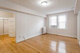 This single-floor residence showcases a practical and well-organized layout spread across several connected rooms. The main living areas are defined by warm, honey-toned hardwood floors that extend throughout, reflecting natural light and lending the space a classic, inviting character. White walls and flush-mount ceiling fixtures keep the aesthetic clean and neutral.

The primary living room is generously proportioned, featuring a cast-iron radiator for heating and a window-mounted air conditioning unit, providing year-round climate control. Multiple windows allow natural light to filter in, brightening the room considerably. An open doorway connects this area to an additional room, also finished with the same hardwood flooring, a radiator, and windows equipped with blinds — suitable for use as a bedroom or secondary living space. A closet with built-in shelving and hooks is accessible from this area.

The galley-style kitchen is efficiently designed, featuring white cabinetry, a gas range, a full-size refrigerator, light stone-toned countertops, and tile flooring. It opens directly toward the living area, maintaining a connected flow.

The full bathroom is finished in light, neutral tones with tile flooring, a white vanity with under-cabinet storage, a mirrored medicine cabinet, a bathtub, and a well-lit vanity fixture above — completing this straightforward and functional residence.