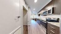 This single-floor unit presents a thoughtfully designed layout that connects key living areas in a clean, contemporary style. The entryway opens directly adjacent to the kitchen, which features dark espresso cabinetry, granite-style countertops, a white tile backsplash, and a full suite of stainless steel appliances including a refrigerator, range, and built-in microwave. Recessed lighting illuminates the galley-style kitchen, which flows seamlessly into the living area. The living room benefits from elevated ceilings, a ceiling fan with a light fixture, and two windows that allow natural light to fill the space. Wood-look plank flooring runs throughout the main living areas, contributing to a cohesive and modern aesthetic. Two bedrooms are carpeted for comfort, each offering a closet with wire shelving and sufficient space for a bed and desk setup. One bedroom includes a window with exterior views. The bathroom is finished with the same dark cabinetry and granite-style countertop found in the kitchen, complemented by a tub-shower combination with grab bar and wood-look tile flooring. Overall, the unit presents a clean, functional, and modern living environment.