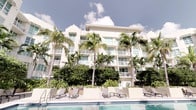 This striking tropical-inspired building presents an elegant white facade that rises across multiple stories, exuding a polished, resort-style aesthetic. The exterior is defined by clean architectural lines, wide horizontal balconies with sleek railings, and an abundance of large windows that allow generous natural light to fill the interior spaces while offering expansive outdoor views.

Towering palm trees line the perimeter of the building, creating a lush, verdant backdrop that enhances the tropical ambiance. The ground level opens onto a well-appointed pool deck, furnished with modern lounge chairs and seating arrangements that invite relaxation. The pool area is thoughtfully designed with manicured landscaping and generous hardscaping, reinforcing the upscale, resort-like character of the property.

Each level features private balconies, providing residents with their own outdoor retreat and the opportunity to enjoy the surrounding tropical environment. The repeating architectural rhythm of balconies and windows across the facade gives the building a cohesive and refined appearance. The overall design balances contemporary elegance with a warm, sun-soaked coastal character, making it a standout example of tropical residential architecture. The combination of lush greenery, open-air amenities, and sophisticated building design creates an inviting and visually impressive living environment.