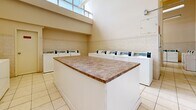 This shared laundry facility offers a generously sized, single-floor space designed for efficient, communal use. The room is defined by its impressively high ceilings, which lend an open, airy quality rarely found in utility spaces. Large clerestory windows positioned near the top of the walls allow abundant natural light to flood the interior, creating a bright and welcoming environment.

The layout is thoughtfully organized, with rows of top-load washing machines and dryers lining the perimeter walls on multiple sides, providing substantial laundry capacity. At the center of the room stands a prominent, island-style folding counter finished with a granite-look laminate surface, offering a practical and spacious area for sorting and folding laundry. A structural column with tiled detailing divides one section of the space, adding an architectural element while maintaining an open feel throughout.

The flooring consists of light-toned ceramic tile laid in a classic grid pattern, contributing to the clean, hygienic aesthetic of the room. The walls are finished with a neutral beige palette and feature a tile wainscoting detail along the lower half, adding a touch of durability and refinement. Fluorescent strip lighting supplements the natural light, ensuring the space remains well-lit at all hours. Informational and operational signage is posted throughout, keeping the facility organized and user-friendly.