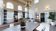 This impressive single-floor residence showcases a grand open-concept layout anchored by a dramatic great room with soaring vaulted ceilings that create an immediate sense of spaciousness and grandeur. Towering arched windows line the walls, flooding the interior with generous natural light while framing views of the surrounding greenery. A striking wrought-iron chandelier hangs as a centerpiece, complementing the warm, neutral tones of the walls and dark hardwood floors throughout.

The living area is thoughtfully appointed with a large tufted sectional sofa dressed in coordinating blue and tan throw pillows, echoing the rich plaid drapery panels that frame each window. A dark wood dining table anchors the foreground, providing a natural transition between the living and entertaining spaces. Wall sconces add a warm, layered lighting ambiance throughout the room.

Adjacent to the main living area, a dedicated workspace or study is visible, outfitted with a desk and shelving, offering a functional retreat without sacrificing the overall cohesive aesthetic. The open sightlines between rooms reinforce the flowing, connected nature of the floor plan. Overall, the space balances elegant architectural details with comfortable, inviting furnishings, resulting in a home that feels both refined and livable.