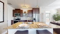 Located on Prairie Winds Drive in St. Charles, Illinois, this single-floor residence offers a thoughtfully designed layout with quality finishes throughout. The kitchen serves as a central highlight, showcasing rich dark-stained cabinetry, a textured mosaic tile backsplash, and warm golden granite countertops extending across a spacious island with an undermount sink. Stainless steel appliances, including a French door refrigerator, slide-in range, and built-in microwave, complement the space, while decorative pendant lighting adds a polished touch. The open layout connects seamlessly to a combined living and dining area, where vaulted ceilings and a row of tall windows allow generous natural light to fill the room. Carpeted flooring runs through the living spaces and bedrooms, lending a comfortable, warm atmosphere. The primary bedroom features crown molding, a large window with outdoor views, and access to a walk-in closet outfitted with multi-tiered wire shelving. The bathroom is finished with wood-look luxury vinyl flooring, a granite-topped dark vanity, and a tiled soaking tub with a built-in niche. A carpeted staircase visible from the main level suggests additional lower-level space.