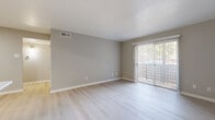 This single-floor residence offers a clean, modern aesthetic highlighted by light-toned hardwood-style flooring that flows consistently throughout the space. The living area is bright and open, featuring a sliding glass door with vertical blinds that provides direct access to an outdoor area and allows natural light to fill the room. A standout feature of the living space is a floor-to-ceiling tiled fireplace with a sleek black metal surround, serving as a refined focal point.

The galley-style kitchen is efficiently designed with gray cabinetry, white countertops, a tile backsplash, and a full suite of black stainless steel appliances including a range, microwave, and refrigerator. A pass-through counter connects the kitchen to the adjacent living and dining area, enhancing the open feel.

One of the bedrooms features a charming bay window with shutters and a ceiling fan, providing excellent natural light and ventilation. This room also includes access to a dedicated walk-in closet with built-in shelving and hanging rods, offering generous storage.

The bathroom is finished with large-format white wall tiles surrounding a soaking tub and shower combination, complemented by a gray vanity and light flooring. Throughout the residence, neutral gray walls and crisp white trim create a cohesive, contemporary atmosphere.