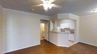 This two-story space showcases a thoughtfully designed open-concept layout connecting the living area and kitchen. The living area features warm hardwood flooring, crisp white crown molding, and a ceiling fan with integrated lighting, contributing to a comfortable and well-lit environment. A peninsula-style kitchen counter extends from the kitchen space, providing a natural division between the two areas while maintaining an open feel. The kitchen is equipped with white cabinetry offering ample storage, a white appliance suite including a refrigerator and range, and countertops that complement the overall neutral color palette. The walls throughout are finished in a soft, neutral tone, creating a clean and versatile backdrop. A doorway visible from the main living area leads to an additional room, suggested to be a bathroom or utility space, with warm lighting. The consistent use of white trim and baseboards ties the spaces together cohesively. The combination of hardwood flooring, crown molding details, and the open layout gives the space a classic yet functional character suited to everyday living across its two floors.