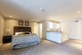 Located at 1547 Mississauga Valley Blvd in Mississauga, Ontario, this single-floor studio suite offers a thoughtfully organized layout that combines living, sleeping, and dining functions within one open space. The main area features soft, patterned carpeting and warm striped wallpaper that lend a cozy, hotel-inspired ambiance throughout. A dedicated seating area near the large window — dressed with vertical blinds — provides an abundance of natural light and a comfortable space for relaxation, complete with cushioned armchairs arranged around a small coffee table. A wall-mounted air conditioning unit ensures year-round climate control. The sleeping area is anchored by a generously sized bed with a dark upholstered headboard, accompanied by framed artwork that adds a refined touch to the walls. The kitchenette is well-appointed with white cabinetry, ample upper and lower storage, a double stainless steel sink, laminate countertops, a microwave, a coffee maker, and a dishwasher — offering full convenience for daily living. Sliding mirrored closet doors contribute to a sense of depth and provide practical storage. The bathroom is finished with neutral tile flooring, a vanity with countertop sink, and a full bathtub with a decorative shower curtain, rounding out this fully functional and self-sufficient suite.