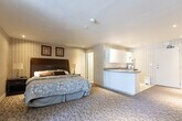 Located at 1547 Mississauga Valley Blvd in Mississauga, Ontario, this single-floor studio suite offers a thoughtfully organized layout that combines living, sleeping, and dining functions within one open space. The main area features soft, patterned carpeting and warm striped wallpaper that lend a cozy, hotel-inspired ambiance throughout. A dedicated seating area near the large window — dressed with vertical blinds — provides an abundance of natural light and a comfortable space for relaxation, complete with cushioned armchairs arranged around a small coffee table. A wall-mounted air conditioning unit ensures year-round climate control. The sleeping area is anchored by a generously sized bed with a dark upholstered headboard, accompanied by framed artwork that adds a refined touch to the walls. The kitchenette is well-appointed with white cabinetry, ample upper and lower storage, a double stainless steel sink, laminate countertops, a microwave, a coffee maker, and a dishwasher — offering full convenience for daily living. Sliding mirrored closet doors contribute to a sense of depth and provide practical storage. The bathroom is finished with neutral tile flooring, a vanity with countertop sink, and a full bathtub with a decorative shower curtain, rounding out this fully functional and self-sufficient suite.