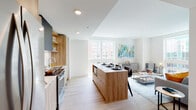 This thoughtfully designed single-floor residence offers a seamless blend of modern aesthetics and functional living. The open-concept kitchen and living area serves as the heart of the home, anchored by a substantial wood-finished island with integrated storage and seating. The kitchen is equipped with stainless steel appliances, including a full-size refrigerator and range, complemented by warm wood cabinetry, open shelving, a herringbone-pattern tile backsplash, and a matte black range hood. Light wood-grain flooring flows throughout, enhancing the bright, airy atmosphere. The living area is bathed in natural light through large floor-to-ceiling windows offering urban views, creating an inviting space with a relaxed yet sophisticated ambiance. The bedroom is a calm retreat featuring a large window for natural light and tasteful décor. The full bathroom is clean and contemporary, with a wide vanity featuring wood cabinetry, a white quartz countertop, a large mirror, and a combination shower/tub with subway tile surround. A hallway with louvered doors provides additional storage and connects the spaces. Recessed lighting throughout ensures consistent illumination, while the neutral white walls provide a versatile canvas across the entire residence.