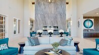 This thoughtfully designed single-floor space presents a striking blend of modern elegance and comfort, unified by a bold teal, grey, and white color palette. The centerpiece of the main lounge is a soaring floor-to-ceiling marble accent wall anchoring a fireplace, framed by exposed wood beam detailing and an industrial-style chandelier overhead. Generous ceiling heights amplify the sense of openness, while large clerestory windows and glass doors flood the interiors with abundant natural light.

The main seating area features plush light-blue upholstered sofas and teal accent chairs arranged around a sleek dark coffee table, complemented by large-scale abstract artwork in oceanic hues. A dedicated lounge nook offers built-in banquette seating upholstered in a teal chevron pattern, paired with round bistro tables and curved accent chairs beneath a series of coordinating geometric wall panels.

A prominent bar area commands attention with a white marble-topped island, barstool seating, and a vivid teal-and-grey chevron mosaic tile feature wall behind it. A separate office or reception area includes a central desk, teal guest chairs, textured wall paneling, and patterned carpeting, creating a professional yet welcoming environment. Light-toned tile flooring flows throughout, tying all zones together seamlessly.