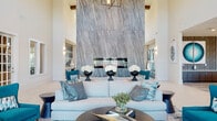 This thoughtfully designed single-floor space presents a striking blend of modern elegance and comfort, unified by a bold teal, grey, and white color palette. The centerpiece of the main lounge is a soaring floor-to-ceiling marble accent wall anchoring a fireplace, framed by exposed wood beam detailing and an industrial-style chandelier overhead. Generous ceiling heights amplify the sense of openness, while large clerestory windows and glass doors flood the interiors with abundant natural light.

The main seating area features plush light-blue upholstered sofas and teal accent chairs arranged around a sleek dark coffee table, complemented by large-scale abstract artwork in oceanic hues. A dedicated lounge nook offers built-in banquette seating upholstered in a teal chevron pattern, paired with round bistro tables and curved accent chairs beneath a series of coordinating geometric wall panels.

A prominent bar area commands attention with a white marble-topped island, barstool seating, and a vivid teal-and-grey chevron mosaic tile feature wall behind it. A separate office or reception area includes a central desk, teal guest chairs, textured wall paneling, and patterned carpeting, creating a professional yet welcoming environment. Light-toned tile flooring flows throughout, tying all zones together seamlessly.