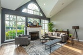 Located at 17600 W 13 Mile Rd in Beverly Hills, Michigan, this single-floor space offers a well-designed, open-concept layout defined by soaring vaulted ceilings that lend an airy, expansive feel throughout. Light hardwood flooring runs continuously across the space, unifying each area and complementing the neutral gray wall palette.

The main living area is thoughtfully arranged with modern gray upholstered seating, including a sofa and coordinating armchairs, centered around a mid-century style round white coffee table. A large, colorful abstract artwork serves as a focal point on the wall, adding a vibrant pop of color. A console table with greenery and a tripod floor lamp contribute warmth and character. Natural light filters in through a large grid-pane window, highlighting the lush indoor plants that animate the space.

The kitchen features dark espresso cabinetry, stainless steel appliances including a full-sized refrigerator and range, and a kitchen island with a marble-style countertop and bar seating. A coffee station is conveniently situated near the entry, enhancing the space's welcoming, functional character. The open sightlines between the kitchen and living areas make the overall layout ideal for both everyday use and hosting.