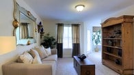 This single-story home offers a thoughtfully designed open-concept floor plan that connects the main living areas for a seamless flow throughout. The living room serves as the heart of the home, featuring plush carpeted flooring, a textured ceiling, and large windows dressed with elegant two-tone drapes that allow generous natural light to fill the space. A decorative mirror, accent shelving, and an armoire contribute to the room's warm, curated aesthetic.

The kitchen is centrally positioned and features a freestanding wooden island, ample cabinetry, and standard appliances, making it both functional and accessible. Adjacent to the kitchen, a dedicated dining area provides a comfortable space for meals and gathering.

The floor plan also reveals a bedroom wing that includes a sleeping area with a ceiling fan and coordinating furnishings. Additional rooms visible in the layout suggest flexible use of space, potentially accommodating a home office or secondary bedroom.

Throughout the home, a consistent palette of warm beige and cream tones creates a cohesive and inviting ambiance. Light-colored carpeting spans the primary living areas, while tiled flooring appears in transitional spaces. Potted plants and tasteful décor accents are present throughout, adding warmth and a lived-in character to the overall environment.