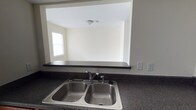 This single-floor space is designed with practicality and openness in mind. The kitchen is a standout feature, highlighted by dark granite-style countertops that provide ample workspace and a double stainless steel sink equipped with a modern faucet. A defining architectural element is the pass-through window cut into the wall above the sink, seamlessly connecting the kitchen to the adjacent living area. This clever design detail promotes an open, airy feel while maintaining a sense of visual connectivity between the two spaces. The living area beyond the pass-through is bathed in natural light courtesy of a window, and is finished with neutral, cream-toned walls that create a clean and welcoming ambiance. A ceiling light fixture in the living area ensures the space remains well-lit throughout the day and into the evening. The overall interior palette is understated and neutral, making it easy to personalize with furnishings and décor. The single-floor layout offers convenient, accessible living with a straightforward and functional floor plan suited to a variety of lifestyles.