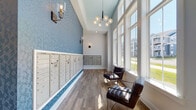 Located at 12525 Oldehurst Place in Charlotte, NC, this single-story mail room is designed with both functionality and aesthetic appeal in mind. The space immediately draws attention with its dramatically high, vaulted ceilings that lend an open, airy quality rarely found in utility-oriented rooms. A wall of oversized, floor-to-ceiling windows lines one side of the room, flooding the interior with generous natural light and offering views of the surrounding community.

The flooring throughout consists of warm-toned hardwood planks that complement the clean white baseboards and neutral wall tones. A striking textured blue accent wall serves as a visual centerpiece, adding depth and character to the space. Rows of individual tenant mailboxes and package lockers are neatly integrated along the walls, providing organized and accessible mail retrieval for residents.

Lighting is carefully considered, with a contemporary globe chandelier suspended from the vaulted ceiling and bronze-toned wall sconces placed throughout, creating an inviting ambiance. Two leather lounge chairs positioned near the windows offer a comfortable waiting area. The overall design balances practicality with a polished, modern aesthetic, making this mail room a refined and welcoming communal space.