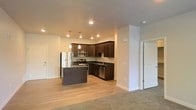 This single-floor residence offers a clean, modern design centered around an open-concept layout that seamlessly connects the kitchen and living areas. The kitchen is a standout feature, showcasing rich dark cabinetry that extends to the upper walls, providing ample storage while creating a bold contrast against the light-toned walls and flooring. A central kitchen island anchors the space, illuminated by stylish pendant lights that add a touch of contemporary elegance. The kitchen is fully equipped with stainless steel appliances, including a refrigerator, microwave, range, and dishwasher, complemented by dark countertops and a stainless steel sink. Recessed lighting throughout ensures the space is well-lit and inviting. Light hardwood-style flooring runs through the kitchen area and transitions into plush carpeting in the living space, creating a comfortable and cohesive flow. The living area is generous and open, offering flexibility for various furniture arrangements. Additional doors visible from the main living area suggest access to further private rooms or storage, completing this well-appointed and functional single-floor residence.