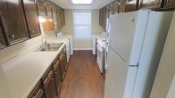 This two-floor residence is designed with everyday functionality in mind, offering a practical and well-organized layout across its levels. The galley-style kitchen is a standout feature, characterized by dark wood cabinetry lining both walls and providing generous storage space. White countertops run the length of the kitchen, complementing the warm hardwood-style flooring beneath. A stainless steel double sink sits beneath upper cabinets on one side, while the opposite wall houses a white electric range and a full-size refrigerator. A skylight overhead fills the kitchen with natural light, enhancing the sense of openness in the narrow but efficient space. A window at the far end, fitted with blinds, adds additional brightness. Notably, the kitchen also accommodates a washer and dryer unit, seamlessly integrating laundry convenience into the home. The overall ambiance of the space is practical and comfortable, with a neutral color palette of beige walls and white trim tying the rooms together. Spread across two floors, the residence offers a clear separation between living and private areas, making it a well-structured home suited for functional daily living.