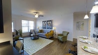 The Blvd At Hayes offers a single-floor residence designed with comfort and style in mind. The living area serves as the centerpiece of the space, showcasing warm wood-style flooring that runs throughout and contributes to a cohesive, inviting atmosphere. Neutral-toned walls provide a versatile backdrop, complemented by crisp white ceilings fitted with a ceiling fan for year-round comfort. Natural light flows generously into the room through multiple windows dressed with blinds, brightening the space throughout the day. The living area is spacious enough to accommodate a full seating arrangement, including a sofa, accent chairs, and a coffee table, all anchored by a patterned area rug that adds visual interest. Decorative wall art and coordinated furnishings enhance the modern aesthetic. The open layout allows for flexible furniture placement and easy flow between areas. Overall, the space presents a clean, contemporary feel with practical finishes and thoughtful design details that make it both functional and visually appealing for everyday living.