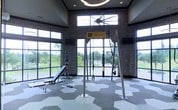 This contemporary fitness center is thoughtfully designed to inspire and support an active lifestyle. The space is anchored by a dedicated TRX Training Zone, complete with a freestanding suspension training rig, making it ideal for functional strength and conditioning workouts. An adjustable weight bench and a rack of free weights provide additional options for resistance training. The flooring features a striking hexagonal tile pattern in neutral tones of grey and white, adding a modern geometric aesthetic underfoot. The walls are adorned with a bold, typography-style graphic wallcovering featuring fitness-related words and phrases, creating an energizing and motivational atmosphere. Elevated ceilings with recessed lighting and a statement circular chandelier contribute to the open, airy feel of the space. Large floor-to-ceiling windows along the perimeter flood the interior with abundant natural light and offer expansive views of the lush green outdoor surroundings, seamlessly connecting the interior environment with nature. Multiple wall-mounted flat-screen televisions are positioned throughout the space for entertainment during workouts. Overall, the fitness center combines function and design in a dynamic, well-lit environment suited for a variety of training styles.