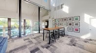 This single-floor clubhouse is designed to foster community and comfort through a blend of modern and transitional design elements. The space is anchored by dramatic vaulted ceilings with exposed beams and industrial ductwork, creating an airy, loft-like atmosphere that feels both open and inviting.

The lounge area is a standout feature, showcasing a full-height exposed brick fireplace that serves as a striking focal point. Built-in bench seating with wood bases lines the large windows, offering comfortable spots flooded with natural light and views of the surrounding outdoors. A tufted leather ottoman coffee table and plush sectional sofa complete the relaxed gathering space, alongside a wall-mounted flat-screen television.

Adjacent to the lounge, a communal dining and work area features a tall wooden table surrounded by modern bar stools, suspended beneath a cluster of pendant lights. A curated gallery wall of framed artwork adds a sophisticated touch. Floor-to-ceiling glass panels open the space visually toward an outdoor pool area, further enhancing the sense of openness.

Throughout, a patterned multi-tone carpet grounds the space while complementing the neutral white walls. The overall ambiance is bright, contemporary, and welcoming — ideal for social gatherings, casual meetings, or relaxed everyday use.