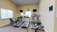This single-floor fitness center is thoughtfully designed to provide a functional and motivating workout environment. The space is outfitted with a solid selection of cardio equipment, including multiple treadmills and elliptical trainers, as well as a stationary bike, catering to a variety of exercise preferences and fitness levels. Wall-mounted mirrors line one side of the room, creating a sense of openness and allowing users to monitor their form during workouts. Two flat-screen TVs are mounted on the walls, positioned for easy viewing from the cardio machines, adding an element of entertainment during exercise sessions. A decorative clock serves as a practical and stylish focal point on the main wall. The space features a drop ceiling with recessed lighting that ensures consistent, even illumination throughout. Neutral-toned walls and a durable, textured floor surface contribute to the room's clean and practical aesthetic. A window with blinds allows for natural light while maintaining privacy. Overall, the fitness center offers a well-organized, comfortable, and fully equipped setting for residents or guests looking to maintain an active lifestyle.