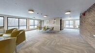 Located at 30 Rockingham Ave in Boston, MA, this single-floor community space is designed to foster gathering, collaboration, and everyday comfort. The layout is thoughtfully organized, offering a spacious open lounge and multipurpose meeting area at its core. Large, floor-to-ceiling windows line the perimeter, flooding the interior with natural light and framing views of surrounding winter trees, creating a bright and welcoming atmosphere throughout.

The main gathering area features herringbone-patterned carpet flooring, warm neutral walls, and a mix of seating arrangements — including yellow-green upholstered chairs grouped around dark tables — suitable for both casual socializing and structured meetings. Colorful framed artwork adorns the walls, adding a lively touch to the otherwise understated décor. A standout feature is an exposed brick accent wall that brings texture and warmth to one section of the space.

A bookshelf stocked with reading materials provides a quiet, resource-rich corner, while a wall-mounted flat-screen television near the entrance offers an entertainment focal point. The space also includes a functional kitchenette equipped with wood cabinetry, a stovetop, oven, microwave, refrigerator, and hardwood-style flooring — supporting everyday practical needs. Overall, the space balances communal functionality with a comfortable, lived-in character.