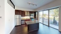 This well-appointed single-floor residence showcases a thoughtfully designed open-concept layout that seamlessly connects the kitchen and living areas. The kitchen is a standout feature, outfitted with rich dark-wood cabinetry, granite countertops, a central island, and a full suite of stainless steel appliances including a refrigerator, electric range, built-in microwave, and dishwasher. Track lighting illuminates the space, while a large sliding glass door opens to an outdoor balcony, flooding the area with natural light and providing outdoor views. The expansive living area continues the open feel with wide-plank dark hardwood flooring, multiple oversized windows framing lush greenery, and additional track lighting overhead. A sliding barn-style door adds a modern architectural touch, separating the living space from the private quarters. The bedroom features neutral carpeting, a ceiling fan with light, and a clean, bright atmosphere. A walk-in closet with wire shelving provides ample organized storage. The full bathroom offers a vanity with a wood cabinet, tiled tub-shower combination, and clean white finishes throughout. The overall ambiance is contemporary, bright, and functional.